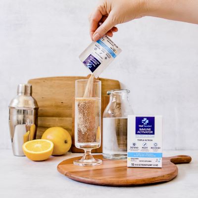A hand pours Well Traveled's Immune Activator into a glass of water placed on a wood board, with lemons in the backround