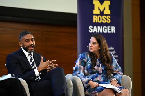 A man and woman speak on stage from gray chairs beside a banner which reads "Sanger"