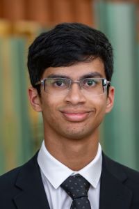 Photo of Michigan Ross student Aayush Sule. He is wearing a black suit jacket with a white shirt underneath with a polka dot black tie. He is wearing glasses and is looking at the camera and smiling.