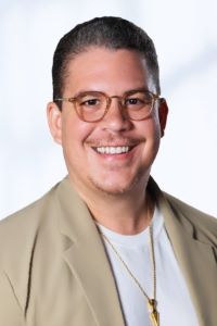 Photo of Executive MBA student Chad Bertrand. He has short dark hair and is wearing brown glasses with a light brown suit jacket with a white shirt underneath with a long gold necklace. He is smiling.