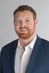 Photo of Michigan Ross Weekend MBA student Chris McCoy who is wearing a navy blue jacket with a white shirt underneath. He has red hair and red facial hair and is smiling. 