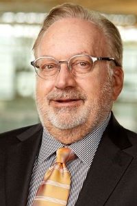 Photo of Douglas Meijer wearing glasses and wearing a black suit and a striped shirt with a yellow tie. 