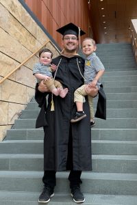 Devin Neu Michigan Ross Executive MBA student is wearing a graduation cap and gown. He is standing on the stairs of the Ross School of Business. He is holding his two young children in both of his arms. 