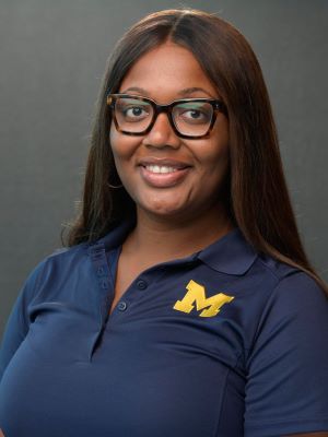 A woman with long brown hair smiles, wearing a blue polo with a yellow block M in front of a gray background