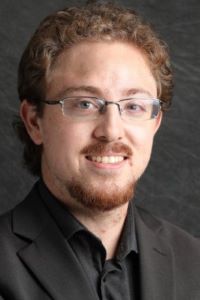 Photo of Michigan Ross Online MBA student Felix Milman. He has red curly hair and facial hair and is wearing glasses and a black jacket with a black shirt underneath. 