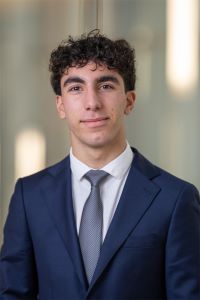 Photo of Michigan Ross BBA student Grant Foumia. He has short dark curly hair and is wearing a navy blue suit with a white shirt underneath and a silver tie. He is looking at the camera and smiling.