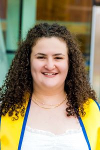 Photo of Michigan Ross BBA student Jessica Goldberg with curly brown hair wearing a white dress with yellow graduation scarf smiling