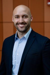 Photo of Michigan Ross Weekend MBA student Jonathan Young who is wearing a navy blue jacket with a light blue shirt. He is bald and has facial hair and is smiling. 