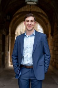 Photo of Michigan Ross BBA student Karim Kadouh wearing a navy blue suit with a light blue shirt underneath. He has dark hair and is smiling. He is standing in front of arched building on the University of Michigan campus.