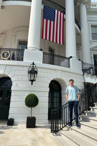 Photo of Michigan Ross BBA student Karim Kadouh standing in front of the Portico steps in Washington D.C. There is an American flag hanging off the building. He is wearing a short sleeve light blue shirt and long pants. 