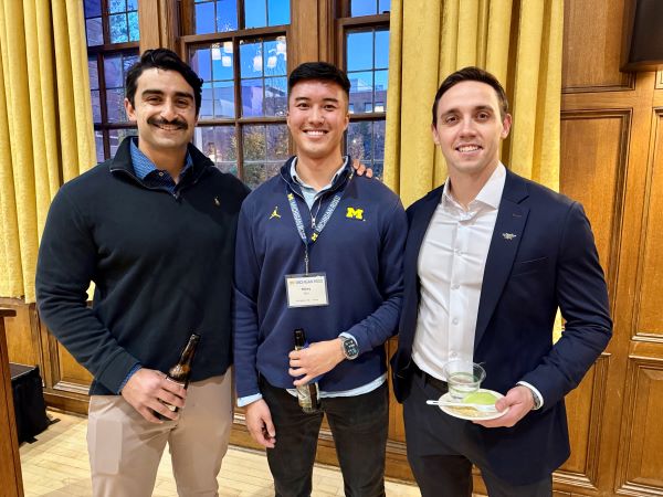Three men wearing Michigan gear smile in a room with wood panel walls