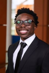 Photo of Michigan Ross BBA student Marvin Tetteh. He has short dark hair and is wearing glasses. He's wearing a black suit jacket with a white button down shirt underneath with a black tie. He is smiling. 