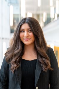 Photo of Michigan Ross BBA student Maya Lindsley, She has long dark wavy hair and is wearing a black suit jacket with a black shirt underneath. She is smiling. 
