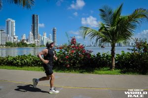 Photo of Merrick Charnet running a marathon in the Great World Race wearing black shorts, black tank top, with buildings in the background and palm trees along the route. 