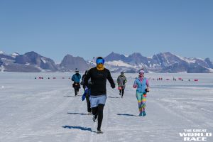 Photo of Merrick Charnet running in the snow in the Great World Race. There are snow covered mountains in the background. He is wearing black tights with blue shorts and long sleeve black shirt. He's also wearing sunglasses and a scarf to keep his face warm.