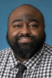 Photo of a man wearing a black and white checkered shirt with a black tie. He has a black beard and mustache and is looking at the camera and smiling.