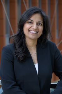 Woman wearing a navy blue jacket with a white shirt underneath. She has long dark hair and is looking at the camera and smiling.
