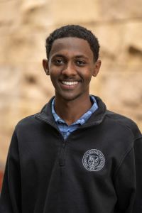 Photo of Michigan Ross BBA student Simon Goitom. He has short dark hair and is wearing a navy blue pullover jacket with a blue plaid shirt underneath. He is smiling. 