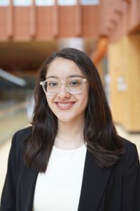 Photo of Michigan Ross BBA student Victoria Guerrero. She has long dark hair and is wearing a black suit jacket with a white shirt underneath and clear glasses. She is standing in the Ross School of Business Wintergarden.