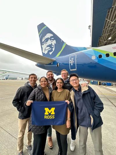 Students hold a Ross flag in front of an Alaska Airlines plane.