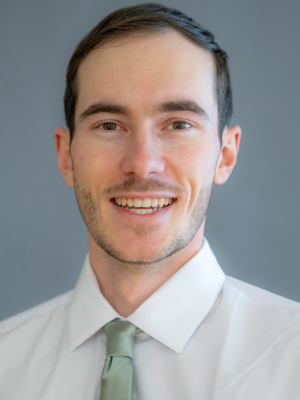 A man with short brown hair smiling wearing a white shirt and green tie in front of a gray-blue background