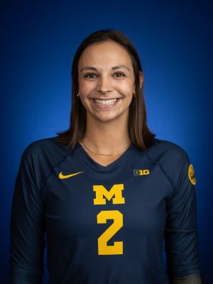 A woman with shoulder length dark brown hair smiling in front of a blue backdrop, wearing a maize and blue jersey with the number 2