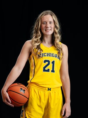 A woman with long blonde hair smiles, holding a basketball and wearing a maize and blue jersey that reads "Michigan #21." She stands in front of a black background