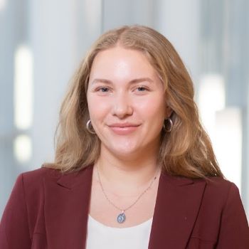 A student with long blonde hair and a maroon blazer with white dress shirt and silver necklace