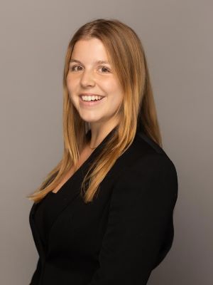 A woman with long blonde hair smiles, wearing a black blazer and shirt in front of a beige background.