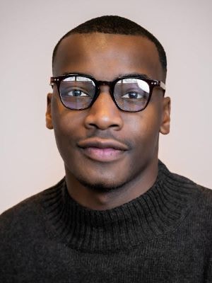 A man with short dark brown hair smiles, wearing glasses and a gray sweater in front of a light gray background