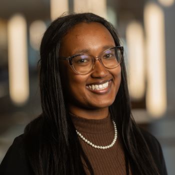 A student with long dark brown hair, glasses, a brown blazer and brown turtleneck with a pearl necklace