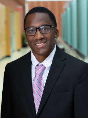 A man with short brown hair and glasses smiles, wearing a black suit and white shirt with pink and blue striped tie. The Ross Winter Garden is in the background