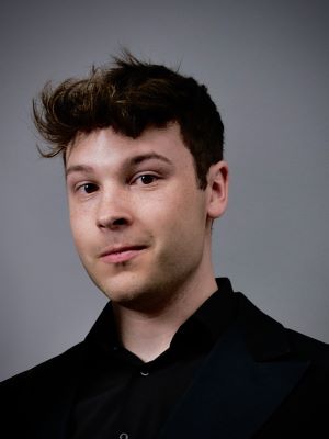 A man with short brown hair posing for a headshot in a black collared shirt, with a gray backdrop