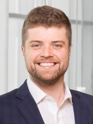 A man with short blonde hair smiles, wearing a blue suit and white dress shirt in a photo taken inside Ross