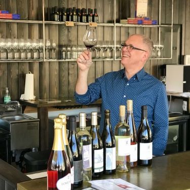 A man holding a wine glass surrounded by wine bottles behind a bar