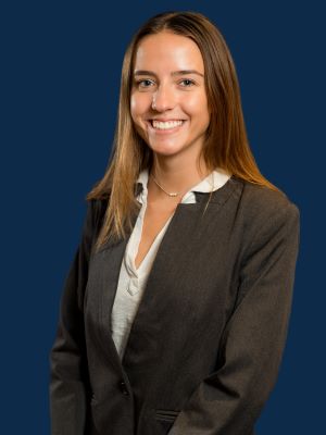 A woman with blonde/copper hair smiling in a gray blazer in front of a blue background