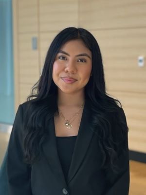 A woman with long black hair smiles, wearing a black blazer and shirt and standing in the Ross building.