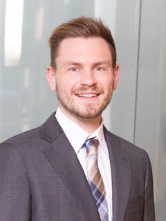 A man with light brown hair wearing a gray suit and plaid tie