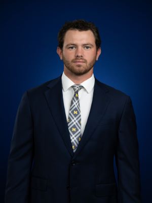 A man with short brown hair poses for a headshot, wearing a suit and standing in front of a dark blue background