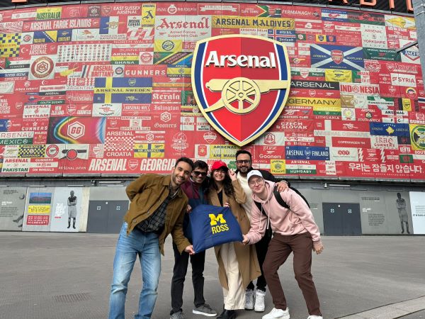 Students stand in front of the colorful Arsenal stadium holding a Ross flag