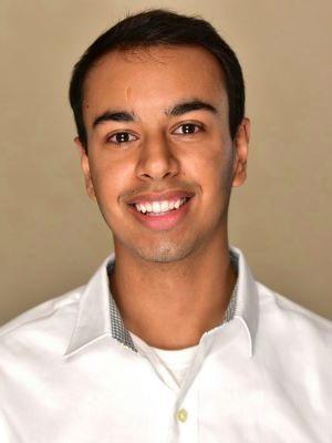 A man with short dark brown hair wearing a white dress shirt smiles in front of a tan background