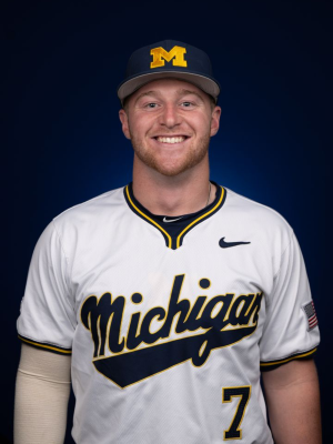 A man with light brown hair smiles, wearing a maize and blue jersey that reads "Michigan #7" and a block M hat in front of a blue background