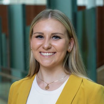 A student with long blonde hair and a yellow blazer with white dress shirt and gold necklace