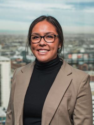 A woman with dark brown hair tied back in an updo smiles, wearing a beige blazer and black turtleneck and standing in front of a cityscape in the window.