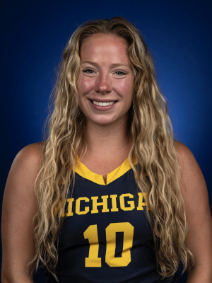 A woman with long, wavy blonde hair in a maize and blue jersey smiles in front of a blue backdrop