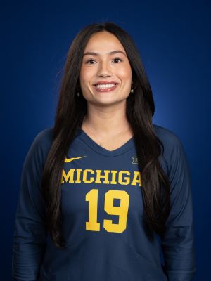 A woman with long brown hair smiles, wearing her maize and blue jersey that reads "Michigan #19"