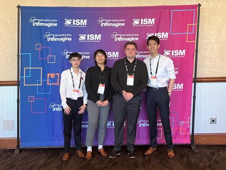 The team stands in front of an ISM banner, each wearing lanyards with access passes for the conference