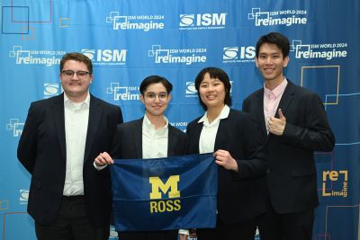 The team stands in front of an ISM banner, holding a Ross flag