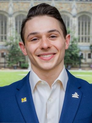 A man with short brown hair smiling, standing in front of the Michigan Law School and wearing a blue suit jacket and white shirt with a block M pin