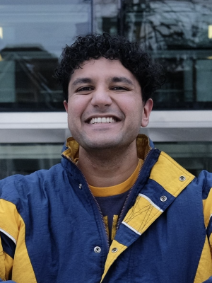 A man with short black hair and a maize and blue jacket smiling in an outdoor area